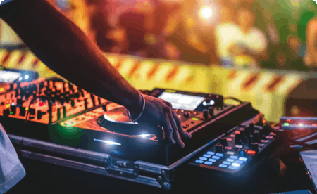 A DJ’s turntable at night, with the DJ’s hand touching it. Blurry people are dancing in the background.